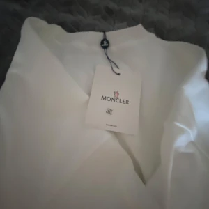 Vit tröja från Moncler - Säljer en stilren vit tröja från Moncler med ett svart emblem på ärmen. Tröjan har långa ärmar och en klassisk passform. Perfekt för en avslappnad och trendig look.