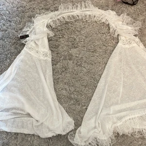 Vit spetsbolero - Elegant vit bolero med spetsdetaljer. Perfekt för att lägga till en romantisk touch till din outfit. Den har en öppen design och är lätt att matcha med olika kläder.