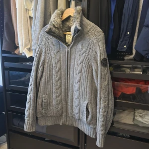 Grå stickad jacka från Gant - Säljer en snygg grå stickad jacka från Gant med kabelstickat mönster och mjukt foder. Jackan har en dragkedja framtill och en hög krage med pälsfoder. Perfekt för kyliga dagar!