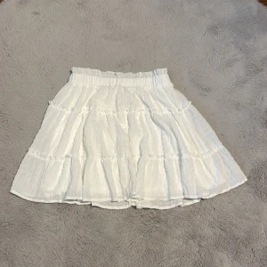 Vit kjol med volanger - Säljer en söt vit kjol med volanger. Kjolen har en elastisk midja och är perfekt för varma dagar. Den är lätt och luftig, vilket gör den bekväm att bära. Passar bra till en enkel topp eller blus.