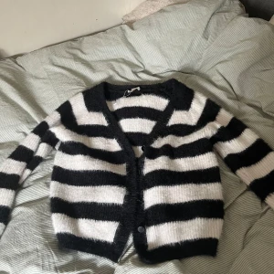 Randig kofta i svart och vit - Mysig och fluffig kofta med breda svarta och vita ränder. Den har en v-ringad design och knappar framtill. Perfekt för att hålla sig varm och stilren.