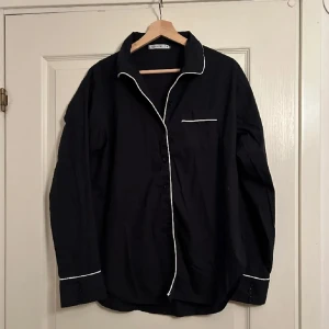 Svart skjorta med vita detaljer - Snygg svart skjorta med vita kontrasterande detaljer längs krage, ärmslut och knappslå. Skjortan har en bröstficka och långa ärmar med knappar vid ärmsluten. Perfekt för en stilren look. Vintage. 