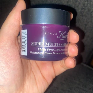 Super Multi-Corrective Cream från Kiehl's - En burk med Super Multi-Corrective Cream från Kiehl's. Denna kräm är designad för att synligt strama upp, lyfta och jämna ut hudens textur. Förpackningen är lila med en silverfärgad lock.