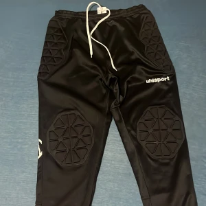 Svarta målvaktsbyxor från Uhlsport - Säljer svarta målvaktsbyxor från Uhlsport med vadderade partier för extra skydd. Byxorna har en elastisk midja med snörning och är perfekta för fotbollsträning. Materialet är syntetiskt och slitstarkt.