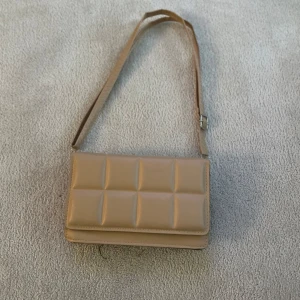 Beige quiltad axelväska - Snygg beige axelväska med quiltad design och justerbart axelband. Väskan har en stilren och modern look som passar perfekt till många outfits. Perfekt storlek för det viktigaste du behöver ha med dig.