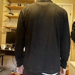 Svart långärmad pikétröja - Säljer en stilren svart långärmad pikétröja med klassisk krage. Perfekt för dig som gillar en enkel och clean look. Tröjan har normal passform och är tillverkad i ett mjukt material som känns skönt mot huden.