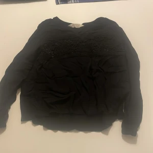 Svart långärmad blus med spetsdetaljer - Säljer en svart långärmad blus med fina spetsdetaljer upptill. Blusen har rund halsringning och är tillverkad i ett tunt, mjukt material. Perfekt att styla med jeans eller kjol.