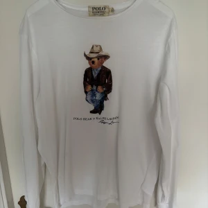 Vit långärmad tröja med Polo Bear från Ralph Lauren - Vit långärmad tröja från Ralph Lauren med det klassiska Polo Bear-motivet på bröstet. Björnen har cowboyhatt, brun jacka och jeans. Tröjan har rund halsringning och är i mjuk bomull. Perfekt för dig som gillar stilrena och lekfulla plagg. Väldigt tunn och skön 