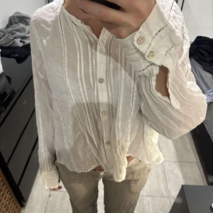 Vit blus med knappar och broderade detaljer - Säljer en luftig vit blus med knappar framtill och diskreta broderade detaljer längs sömmarna. Blusen har lång ärm med manschett och en avslappnad passform. Perfekt för dig som gillar en stilren och enkel look.