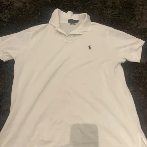 Vit piké från Polo Ralph Lauren - Klassisk vit pikétröja från Polo Ralph Lauren med broderad logga på bröstet. Tröjan har krage och korta ärmar, tillverkad i bomull för en bekväm känsla. Perfekt för en stilren och tidlös look.