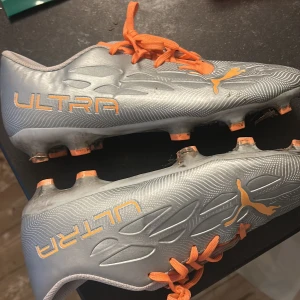 Puma Ultra fotbollsskor i silver och orange - Säljer ett par Puma Ultra fotbollsskor i silver med orange detaljer och snörning. Skorna har en smal och sportig design med rund tå och platt sula, perfekta för snabba rörelser på planen.