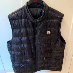 Svart dunväst från Moncler - Svart dunväst från Moncler med quiltad design och dragkedja framtill. Västen har två fickor med dragkedja och Moncler-logga på bröstet. 