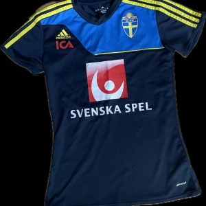 Svenska landslagets fotbollströja från Adidas - Mörkblå fotbollströja med gula detaljer och Svenska landslagets emblem på bröstet. Adidas-logga och ICA-tryck på framsidan, samt stor röd Svenska Spel-logga både fram och bak. Kortärmad modell med sportig passform och ventilerande material.