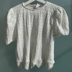 Vit spetsblus med puffärm - Säljer en söt vit blus med kort puffärm och vackra spetsdetaljer. Blusen har rund halsringning och dekorativt mönster i tyget samt vågig nederkant. Perfekt för dig som gillar romantisk stil!