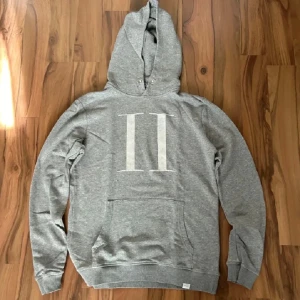 Grå hoodie från Les Deux - Säljer en grå hoodie från Les Deux med stor vit logga på bröstet. Tröjan har huva och magficka, perfekt för en avslappnad stil. Klassisk passform och mjukt material.