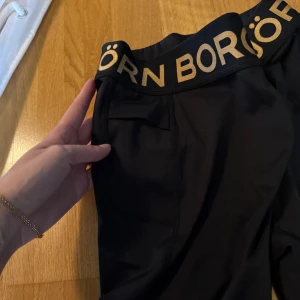 Svarta tights från Björn Borg med guldtext - Svarta tights från Björn Borg med bred resår i midjan och guldig logotyp. Tightsen har dekorativa detaljer med små prickar vid bensluten. Perfekta för träning eller en sportig look. Passar även small