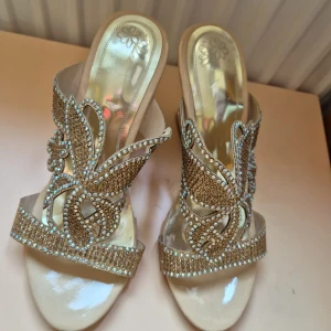 Guldiga klackskor med strass och fjärilsdetalj - Säljer ett par glittriga guldiga klackskor med öppen tå och dekorativa fjärilar i strass. Skorna har en hög klack med utskurna blommönster på sidan och bred rem framtill. Perfekta för dig som vill sticka ut med extra bling. Inte använda