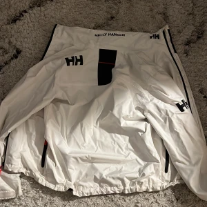 Vindjacka Helly Hansen stl. S - Säljer en snygg vit vindjacka från Helly Hansen med svarta och röda detaljer. Jackan har dragkedjor på fickorna, hög krage och meshfoder på insidan. Perfekt för dig som gillar stilren och sportig look. Nästan aldrig använd. Säljer för den ej passar längre. Passar folk runt 170-185 beroende på din vikt. 