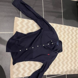 Mörkblå skjorta från Ralph Lauren - Klassisk mörkblå skjorta från Ralph Lauren med vit knappstängning och röd broderad logga på bröstet. Skjortan har lång ärm och en tidlös design som passar till många tillfällen.