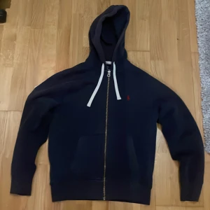 Mörkblå hoodie med dragkedja från Polo Ralph Lauren - Pris går att diskuteras! Säljer en mörkblå hoodie från Polo Ralph Lauren med dragkedja framtill och vit snörning i huvan. Tröjan har fickor på magen och en liten röd logga på bröstet. Perfekt för en avslappnad stil. Tröjan är gott skick och inget fel på!