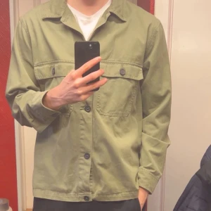 Olivgrön overshirt med knappar - Säljer en stilren olivgrön overshirt med knappar framtill. Jackan har två bröstfickor med knappar och långa ärmar. Perfekt för lager-på-lager-stil. Passar både till vardag och mer avslappnade tillfällen.