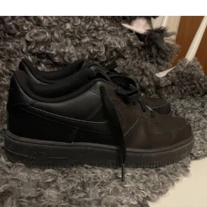 Svarta Nike Air Force 1 sneakers - Klassiska helsvarta Nike Air Force 1 sneakers med låg profil. Skorna har svart snörning, rund tå och är tillverkade i skinn med syntetdetaljer. Perfekta för en clean och stilren look. Väldigt sparsamt använda. Vill få sålda då de ej används.