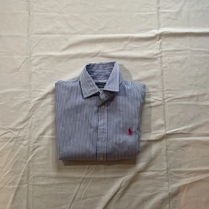 Blåvit randig skjorta från Polo Ralph Lauren - Snygg blå och vit randig skjorta från Polo Ralph Lauren med klassisk krage och knappar framtill. Skjortan har lång ärm och den ikoniska röda broderade loggan på bröstet. Perfekt för en stilren look.