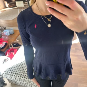 Mörkblå långärmad topp med volang från Ralph Lauren - Säljer en mörkblå långärmad topp från Ralph Lauren med volang nertill och den klassiska rosa loggan broderad på bröstet. Toppens material känns mjukt och stretchigt, storleken sitter mer som en S