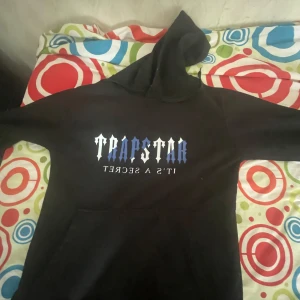 Svart hoodie från Trapstar - Säljer en svart hoodie från Trapstar med tryck i blått och vitt på bröstet där det står 'TRAPSTAR IT'S A SECRET'. Klassisk huva och känguruficka framtill. Perfekt för en avslappnad streetwear-look.