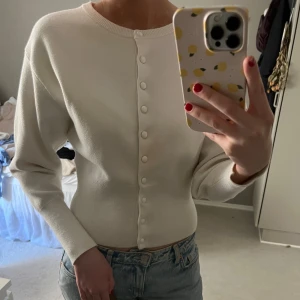 Vit ribbad kofta med knappar - Säljer en stilren vit kofta med ribbad struktur och knappar framtill. Tröjan har lång ärm och sitter figurnära, perfekt att matcha med jeans eller kjol.