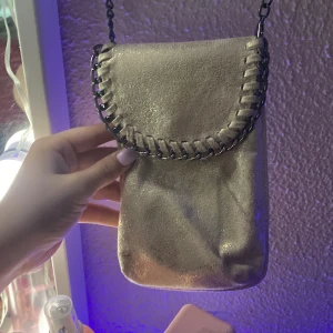 Guldig axelväska med kedjedetalj - Snygg liten axelväska i guldigt material med lock och dekorativ kedja runt kanten. Perfekt för att bära mobilen och småsaker. Väskan har en lång kedjerem och stängs med tryckknapp.