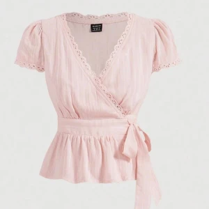 Ljusrosa omlottblus med spetsdetaljer - Supersöt ljusrosa omlottblus med knyt i midjan och spets längs v-ringningen. Blusen har korta ärmar med volang och ett lätt, luftigt tyg med diskreta ränder. Perfekt för sommaren.