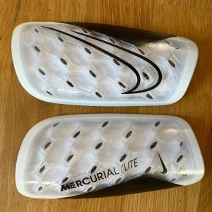 Nike Mercurial Lite benskydd - Säljer ett par Nike Mercurial Lite benskydd i vitt och svart. De har en lätt och smidig design och mjuk insida för extra komfort. Perfekta för fotbollsträning och match. Levereras med originalpåse. Andvänd 1 gång på grund av att de var för stora. 150-160