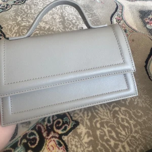 Ljusgrå handväska från Ginatricot - Säljer en stilren ljusgrå handväska från Ginatricot med enkel design och magnetlås. Väskan har ett handtag upptill och ett innerfack med dragkedja. Perfekt för att bära det viktigaste med stil.