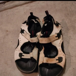Beige och svarta sandaler med kardborreband - Säljer ett par sportiga sandaler i beige och svart med justerbara kardborreband. Skorna har öppen tå och platt sula, perfekta för varma dagar och utomhusaktiviteter. Snygg och praktisk design med svarta detaljer.