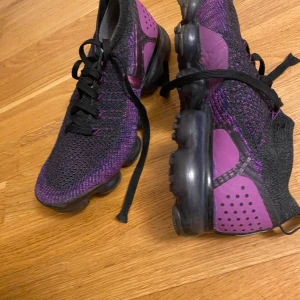 Nike Air Vapormax Flyknit sneakers i lila och svart - Säljer ett par Nike Air Vapormax Flyknit sneakers i lila och svart. Skorna har svart snörning, stickad ovandel och genomskinlig sula med bubblor. Perfekta för dig som gillar sportig stil och vill ha något unikt.