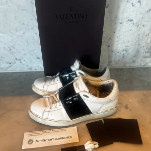 Valentino opens  - Säljer ett par vita Valentino Garavani Open sneakers med bred svart läderdetalj över mitten. Skorna har snörning och rund tå, samt subtila nitar på hälen. Klassisk och stilren design med märkets logga på plösen. 41-42