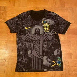 Svart Brazil tröja med Jesusstatyn - Unik svart Brasilien tröja från Nike med stort tryck av Kristusstatyn och detaljerade mönster. Det är en väldigt skön, avslappnad och stilig tröja och nu perfekt till sommaren. Den är i väldigt bra skick har bara testat vid några tillfällen. Priset kan diskuteras vid snabb äffär. Vid frågor är det bara fråga:)