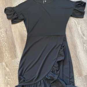 Svart klänning med volang från Vero Moda - Svart klänning från Vero Moda med korta volangärmar och omlottvolang nertill. Mjuk och stretchig i materialet. Rund halsringning och normal passform. Använd fåtal gånger. 