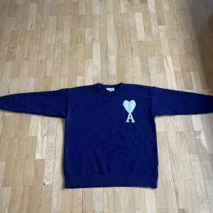 Mörkblå stickad tröja från Ami Paris  - Säljer en mörkblå stickad tröja från Ami Alexandre Mattiussi med vit hjärt- och A-detalj på bröstet. Klassisk rund halsringning och ribbade muddar vid ärmslut och nederkant. Perfekt för dig som gillar stilrena plagg med en twist.