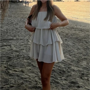 Beige volangklänning med tunna axelband - Säljer en superfin beige kortklänning med flera lager volanger och tunna axelband. Klänningen har en luftig och flowy passform, perfekt för varma dagar eller sommarens tillfällen. Stilren och enkel att matcha med accessoarer.