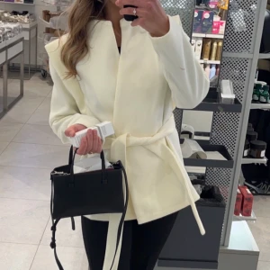 Vit kappa med bälte - Snygg vit kappa med knytbälte i midjan och stilren design. Jackan har lång ärm och rak passform, perfekt för dig som gillar en clean look. Passar till många olika outfits och är lätt att matcha.