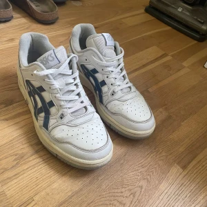 Vita Asics skor stl 44 - Säljer ett par vita Asics sneakers med blå ränder på sidorna. Skorna har rund tå, snörning och perforerade detaljer framtill för extra ventilation. Klassisk sportig look med platt sula och diskret Asics-logga på plösen. Smått slitage, knappt synligt, hör av dig vid frågor eller om du vill ha fler bilder😊