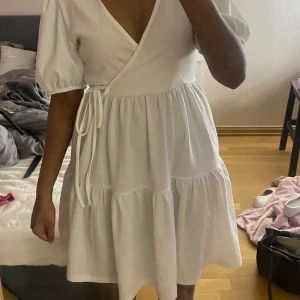 Vit omlottklänning från Gina Tricot - Superfin vit omlottklänning från Gina Tricot med puffärmar och knyt i midjan. Klänningen har en v-ringad hals och volangdetalj nertill. Perfekt för sommarens alla tillfällen! Bara andvänd en gång så som ny💕
