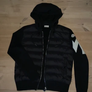 Svart Moncler cardigan med huva - Säljer en svart cardigan från Moncler med huva och dragkedja. Jackan har stickade ärmar med ett vitt sicksackmönster på vänster ärm och Moncler-logga på axeln. Framsidan är quiltad och vadderad för extra värme.