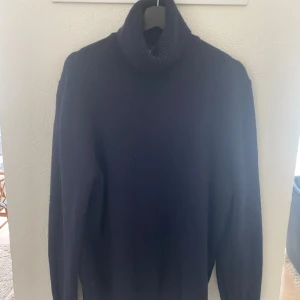 Mörkblå stickad polotröja från H&M - Säljer en mörkblå stickad polotröja från H&M med regular fit. Tröjan har en hög, ribbad polokrage och långa ärmar. Perfekt för kyliga dagar och enkel att matcha med olika outfits.