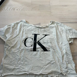 Vit t-shirt från Calvin Klein Jeans - Vit t-shirt från Calvin Klein Jeans med stor svart CK-logga framtill. Klassisk rund halsringning och korta ärmar. Croppad. Perfekt för en enkel och stilren look.