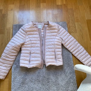 Ljusrosa quiltad pufferjacka - Säljer en ljusrosa quiltad pufferjacka med lång ärm och dragkedja framtill. Jackan har en smal passform och dekorativa band längs dragkedjan. Perfekt för vår och höst.