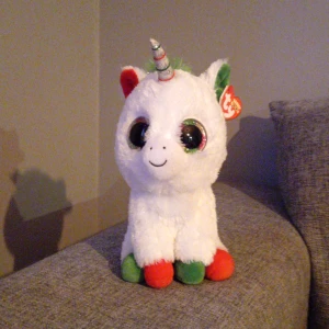 Vit enhörning gosedjur från Ty - Supergulligt gosedjur i form av en vit enhörning från Ty med stora glittriga ögon, röd och grön man samt silverfärgat horn. Fötterna är röda och gröna. Perfekt för dig som älskar mjukdjur och magiska varelser.