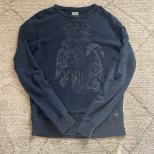 Tröja från Denim & Supply Ralph Lauren - Säljer en mörkblå sweatshirt från Denim & Supply Ralph Lauren med ett stort, slitet örntryck på bröstet. Tröjan har rund halsringning och långa ärmar. Perfekt för en avslappnad stil.